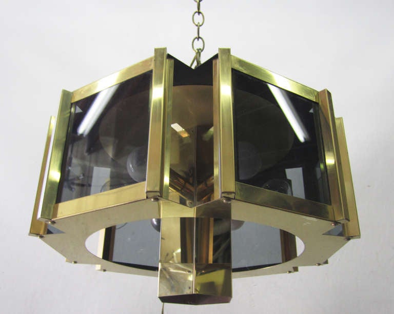 This glamorous brass chandelier by Fredrick Ramond  features six smoked glass panels each paired with an outward-facing bulb. The geometric theme carries over to the central column which extends above and below the glass panels and is fitted with a