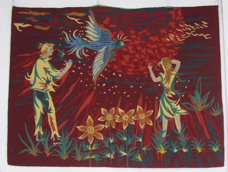 This very romantic Mid-Century Modern tapestry from France depicts a young couple and a large blue bird against the background of a fiery sky. The figures stand in a field of flowers and plants as they watch the creature take flight perhaps to join