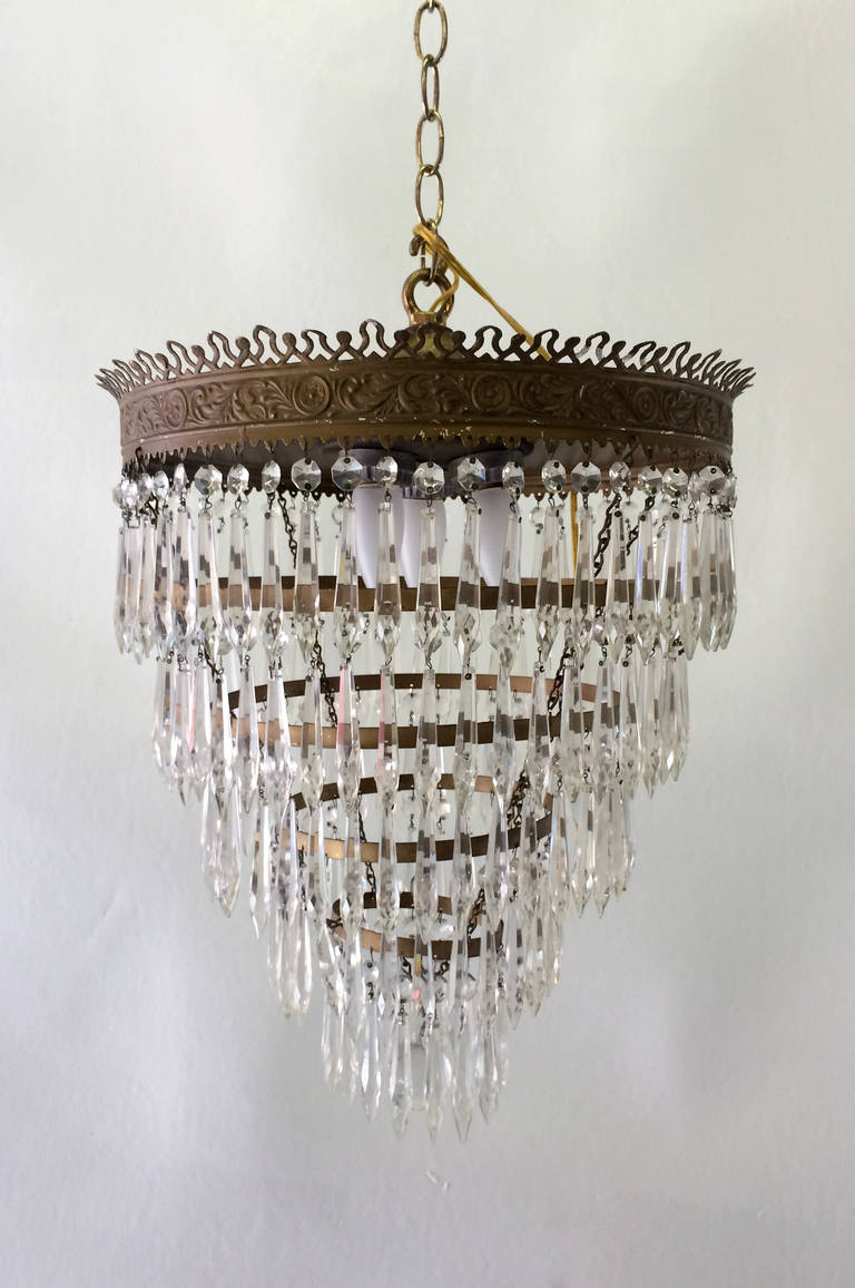 This glass chandelier features a five-tier brass frame in an inverted wedding cake shape. The uppermost ring of brass has a beautiful filigree motif that looks like a crown. The five brass tiers are decorated with dripping tear-shaped glass dangles,