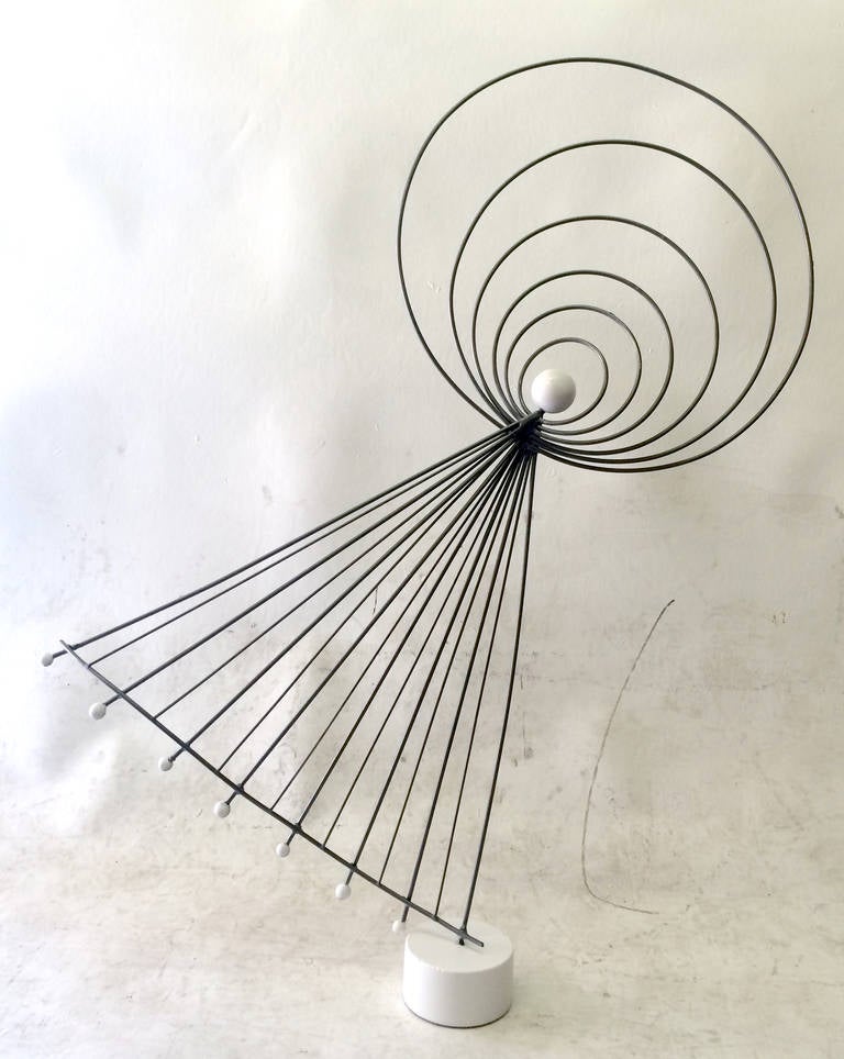 A unique sculpture composed of metal rings and rods welded at the center to create a sort of humanoid floating shape. This piece is weighted at the center with a metal sphere and anchored by a heavy circular metal base. The seemingly weightless
