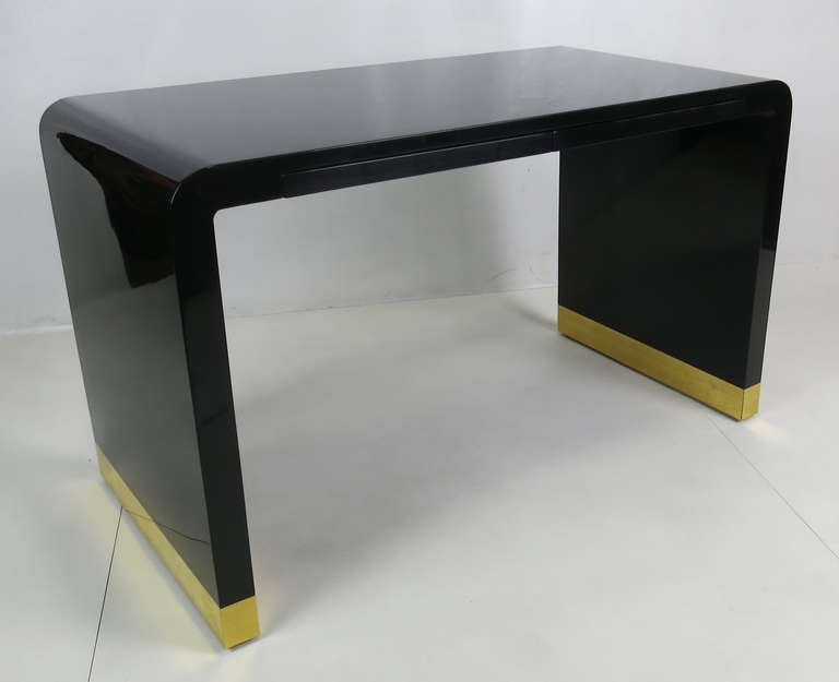 Glamorous 80's Waterfall Writing Desk freshly refinished in French Polished Black Lacquer.  The base has also been freshly restored in heavy gauge solid polished brass.  Two concealed pencil drawers in apron.