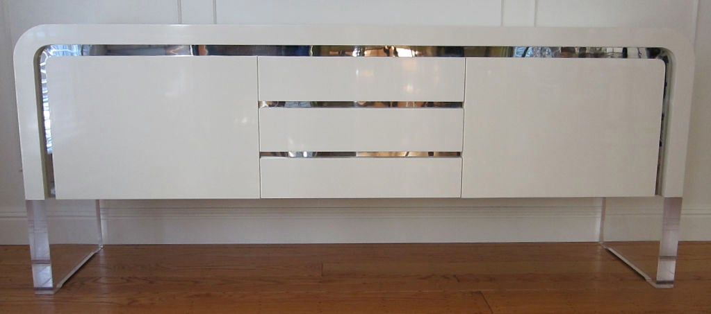 Waterfall Credenza or Buffet with chrome trim and Slab Lucite Legs by Vladimir Kagan.  The piece features three drawers and two doors concealing adjustable shelves.  Meticulously restored in Bone White Lacquer.
