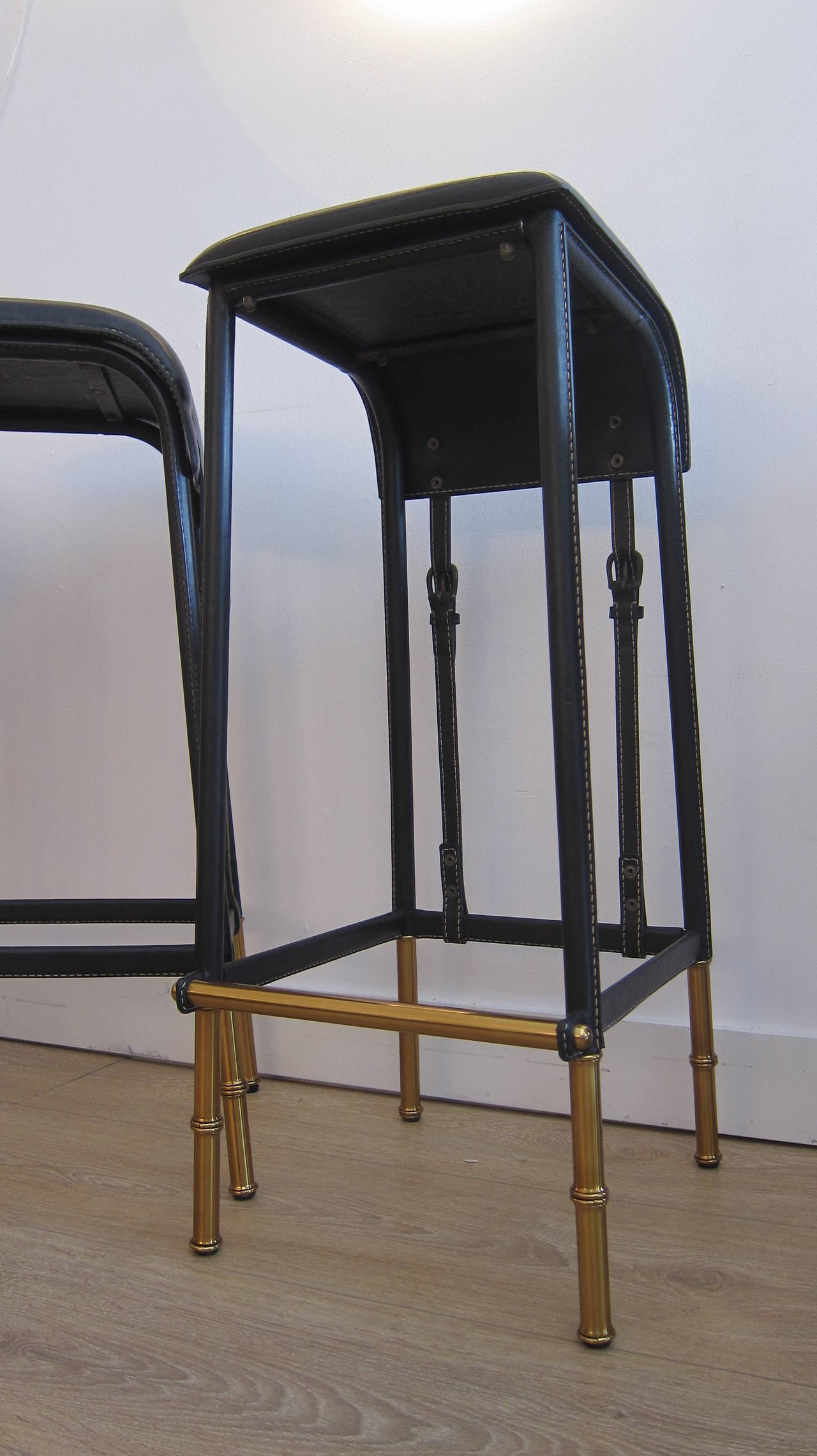 A pair of black stitched leather, saddle bar stools by Jacques Adnet. Brass faux bamboo legs. Four available.
