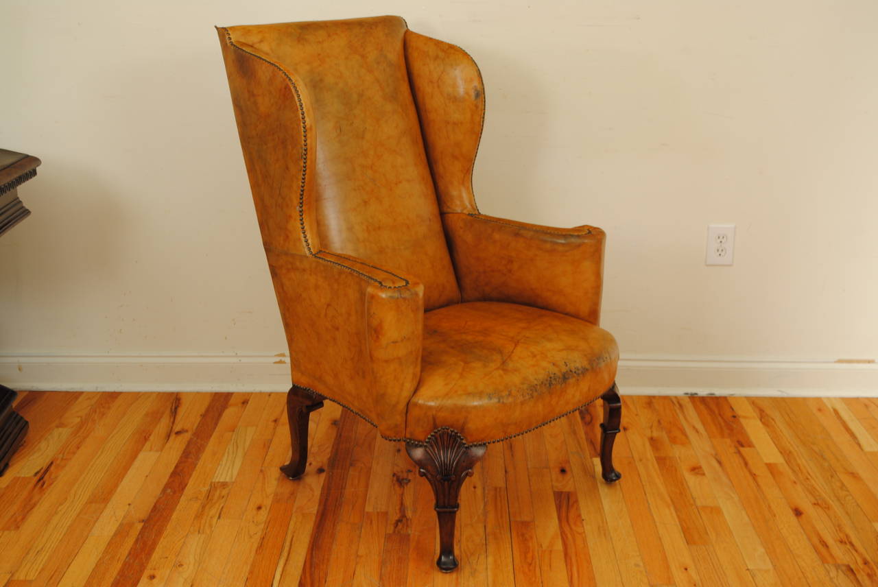 The wingback covered in tan leather trimmed in nailheads, the front legs with shell carvings, the back legs splayed backwards.