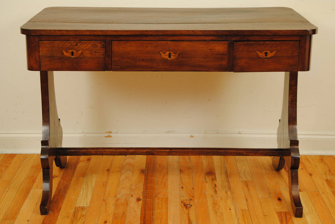 The rectangular top having rounded edges and housing three drawers with inlaid escutcheons, raised on trestle-form legs and joined by a straight stretcher.