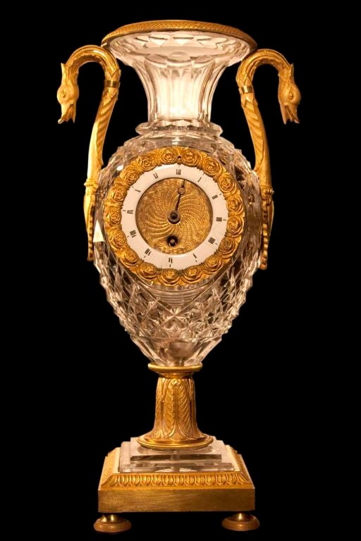 # V139 - Elegant Empire cut crystal urn vase clock enriched with gilded bronze details including chased rim, pair of attenuated swan-neck handles, column, and square gilded bronze base with engine-turned feet.
French, Circa 1815
RESTORATIONS: