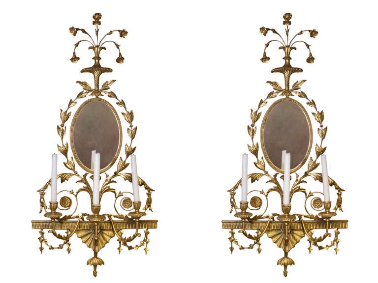 # S094 - PAIR George III carved and giltwood wall sconces in the manner of Robert Adam. The tops surmounted by neoclassical urns with a spray of flowers above an oval mirror plate surrounded by carved borders and foliage. A fluted crosspiece with