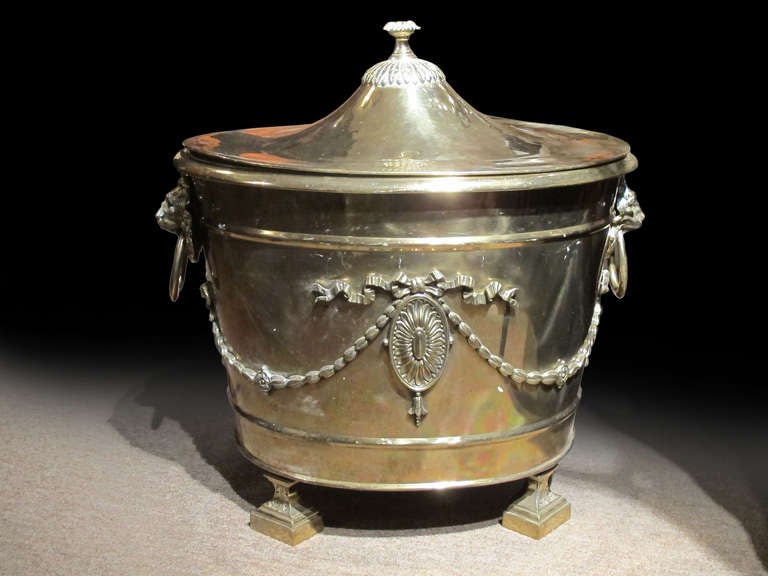 # Y029 - Victorian brass bucket and cover in the Adam taste. The pure neoclassical vocabulary of motifs compliments the oval shape. Note the draped bell flowers from the pair of lion head handles meeting at the oval paterae. The cover with a foliate