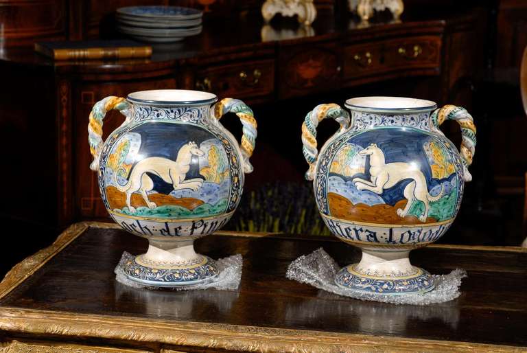 Pair of twisted, double handled, neoclassical, porcelain vases. Blue is the primary color surrounding the proud dogs on their hind legs but, you also see browns, golds and greens sprinkled throughout to compliment any color pallet.