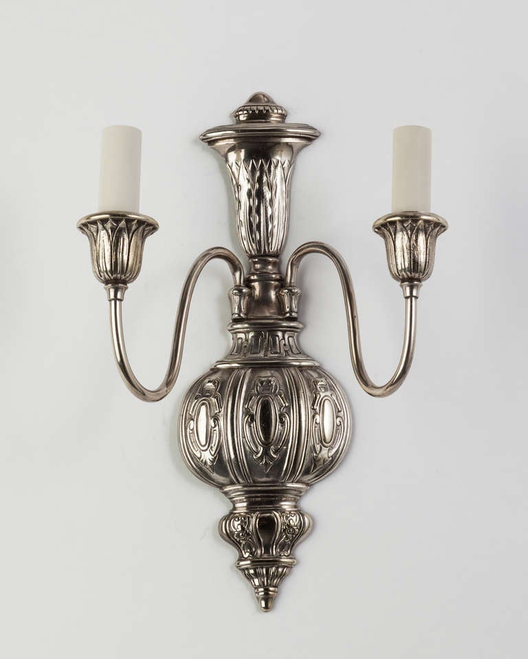 AIS2870
A pair of two arm silverplate over cast bronze sconces. The backplates are carved in low relief with cartouches and foliate details; the cups wrapped with a leaf and dart motif. Circa 1920s.

Dimensions:
Overall: 13-1/2