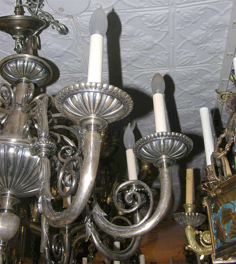 Lampadario a otto luci in stile neoclassico in bronzo argentato con finitura e patina originali. I bracci sono caratterizzati da un motivo a volute e a fogliame.