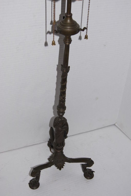 A cast bronze neoclassic style tripod base table lamp, with figure on body and tripod base. Foliage details on body.
Measures:
24