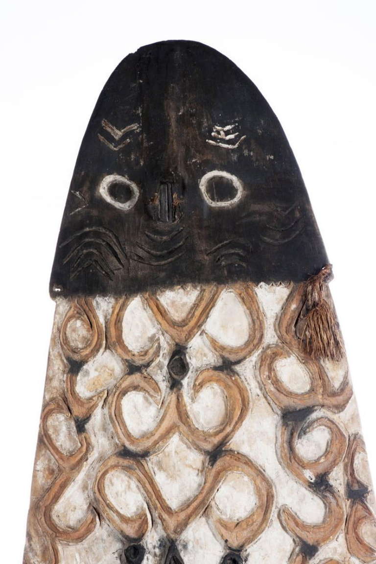 From Western Asmat territory, the shield with raised curvilinear designs in red and black against a white ground. The top with stylized head of a turtle or rayfish. The back with large handle, scribbles all over the back. A fine example on a custom