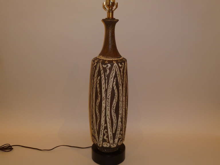 With a wonderful textured brown lava like glaze punctuated with seemingly etched designs of squigglies and snaking figures in white, this Italian pottery lamp is in the style of Gambone. Standing on an ebonized circular wood socle base, the table