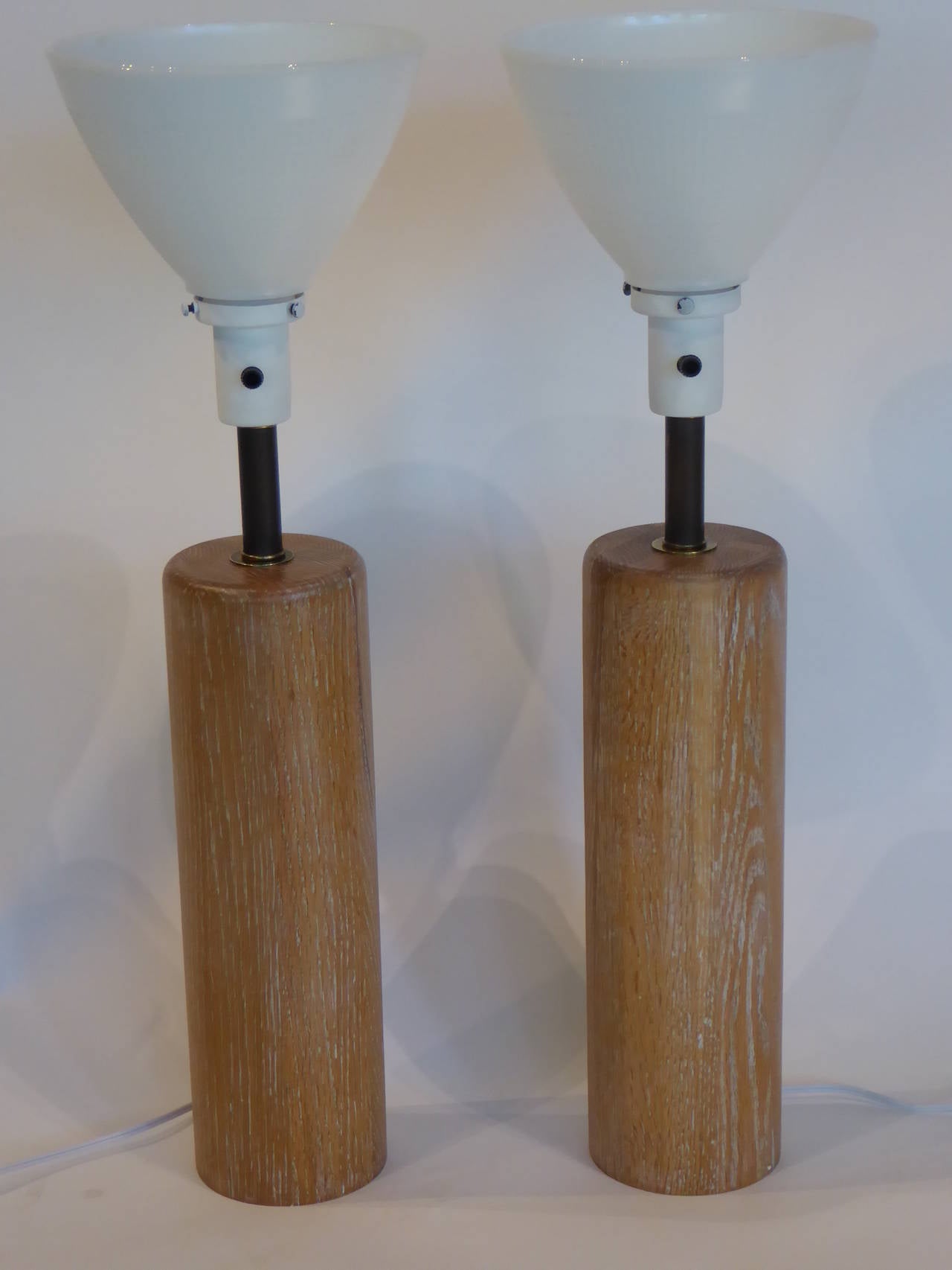 Beautiful figured cerused or limed oak highlights this pair of Nessen Studios table lamps. Classic and timeless, they are comfortable in all modern era styles. Rich wood columns rise to a bronze patina brass neck to the milk glass torchiere shade