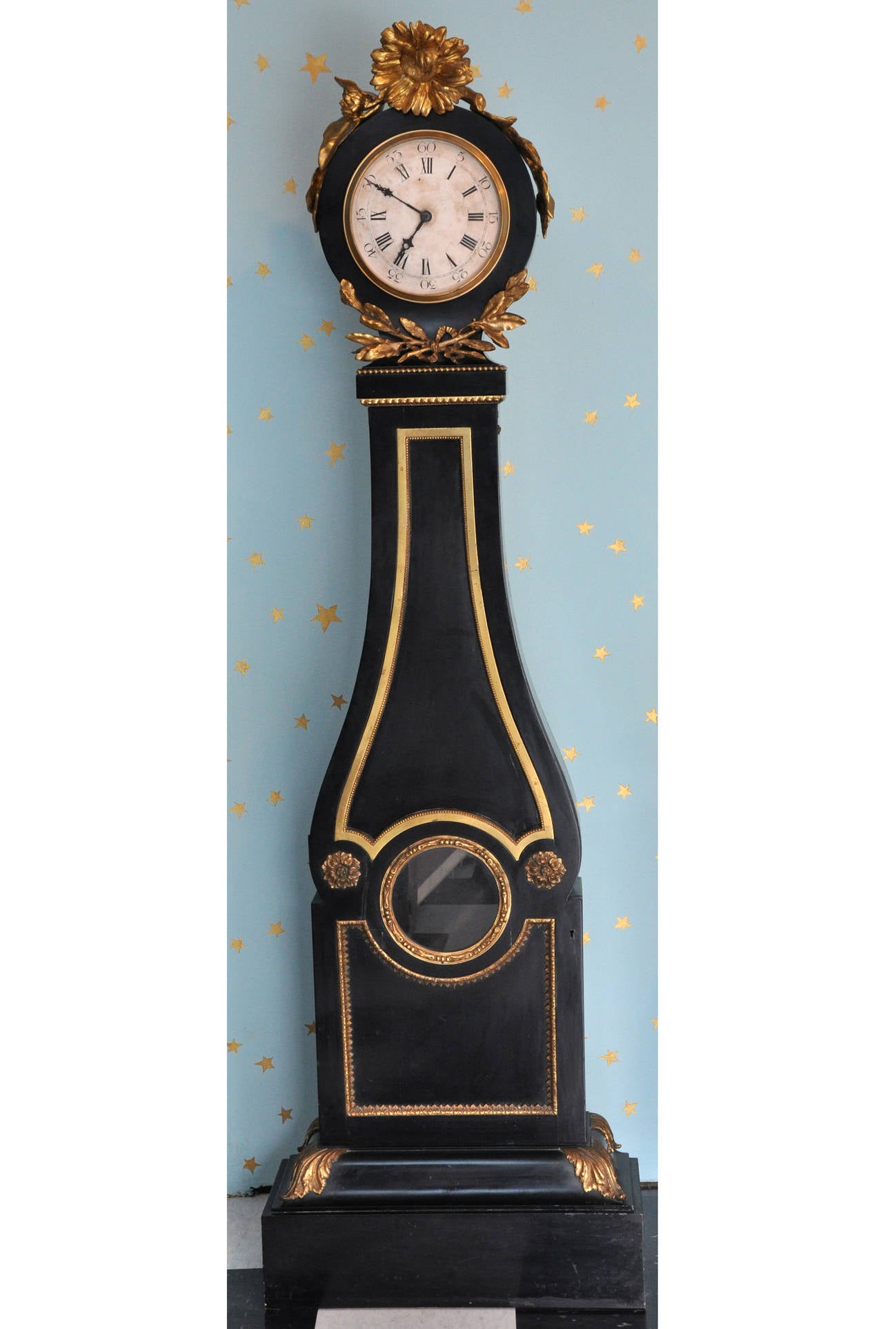 Antique French tall case clock - vintage 19th Century.  Clock has been converted to battery operated.  Clock has applied bronze wreath decorations and the top of the clock is crowned with a beautiful gold leafed carved wood flower with trailing