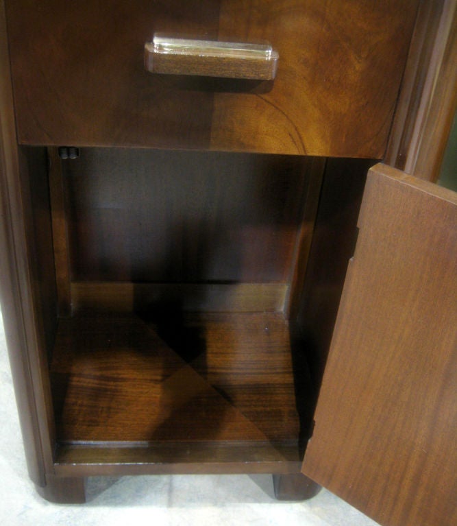 Donald Deskey (1894 – 1989) designed this American art deco bedside table for his own company AMODEC (American Modern Decoration Company) in 1931.  The stand, in figured walnut veneers, has been refinished and the metal pulls on the drawer and door