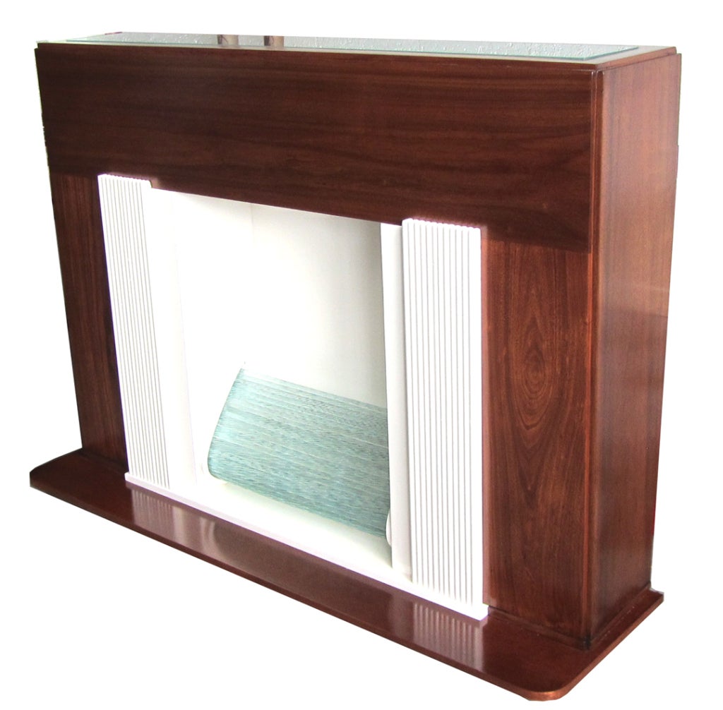 This sleek American art deco electric fireplace is from the 1930’s. The cabinet is mahogany and has a frosted glass inserted top. The “fire” is in a space flanked by ribbed pillars all in white lacquer. The “fire” is twisted rods glass, illuminated