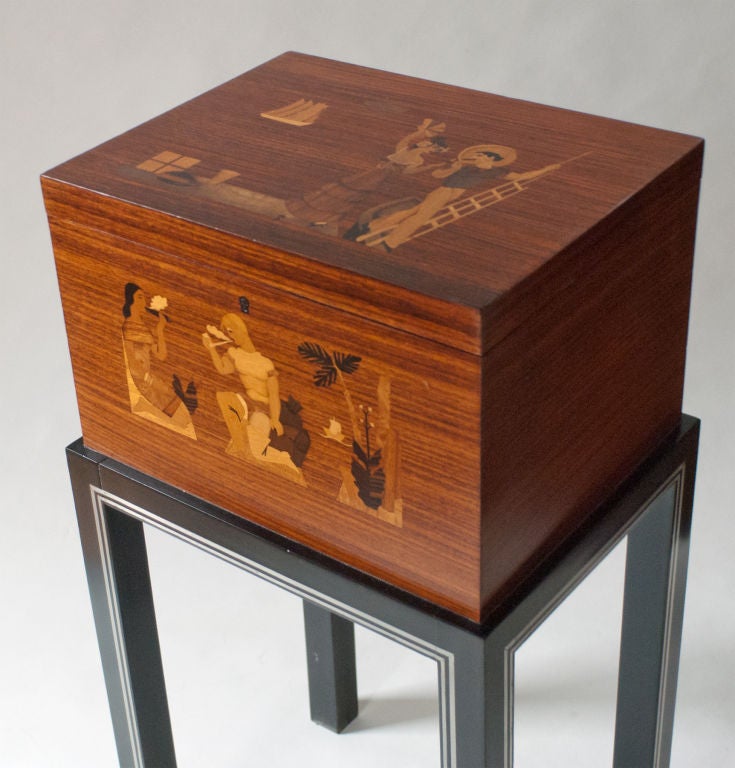 Swedish Art Deco lidded chest on four legs in mahogany with two marquetry scenes. Front depicts a native American and a European explorer smoking with a tobacco plant and a ship chest besides them. The lid has a Cuban couple on a dock with a woman