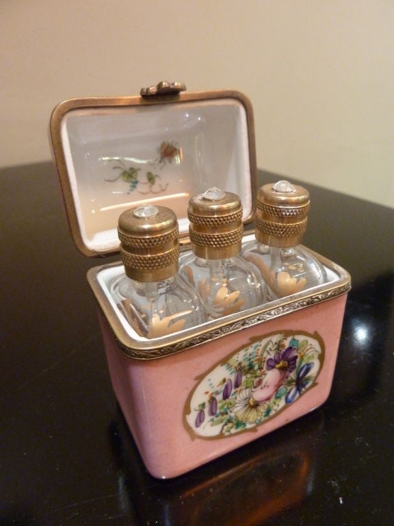 A magnificent Limoges box is artfully painted in cheerful pink with floral art on the front façade and top lid.  Inside are three gold painted glass perfume bottles with jeweled caps. The box is adorned with intricate metalwork and an ornate flower
