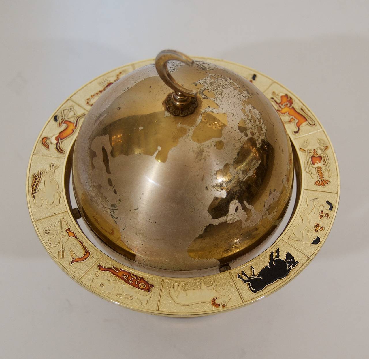 Unusual Mid-Century cigarette holder, the upper hemisphere extending to reveal cigarette storage. Surrounding by an unusual enameled brass ring with the signs of the Zodiac. Brass, enamel and nickel plating.