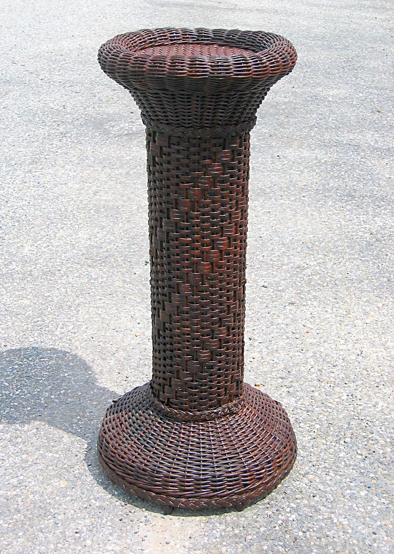 Victorian wicker pedestal in faux-natural stained finish. Serpentine rolled edge on top with basket-woven flat surface. 
Decorative repeating spiral pattern woven on cylinder. Bulbous round base having turned wooden ball feet. Made of reed.