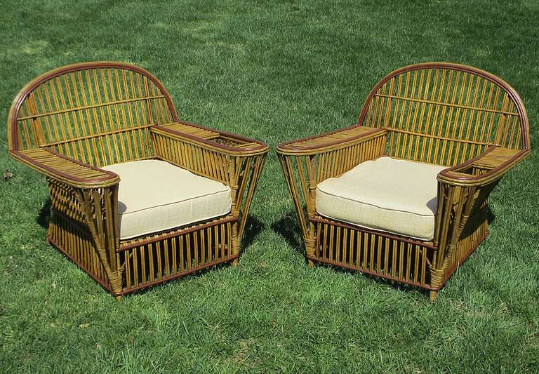 Matching pair of large scale Stick Wicker armchairs in original honey-toned color with burgundy painted trim.  Deep-seated, wide flat arms and  magazine pockets woven into the left side of both chairs.  Unusual cone shaped facade to arms.  Closely