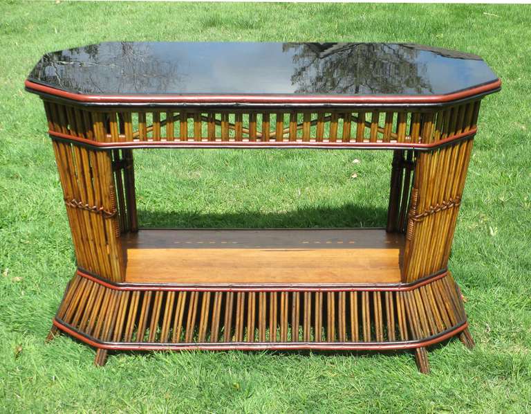 Stick Wicker console table with black glass table top.  Natural stained finish with red & black painted highlighting.  Octagonal at ends, large lower bottom wood shelf, close vertical paired reeds, supported on 8 legs.