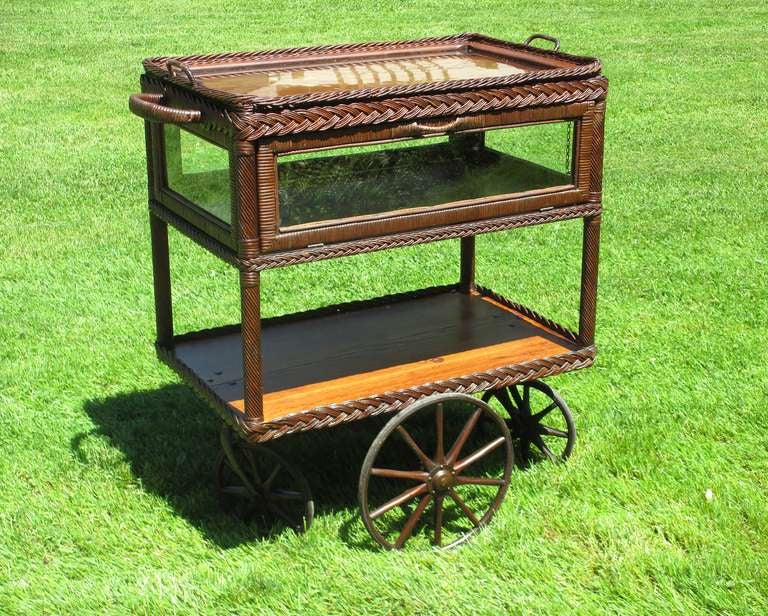 Very unusual wicker tea cart having 4-sided glass enclosed pie safe component with 2 drop down doors. Sold oak surfaces and woven reed & cane in original natural stained finish. Cane-wrapped handles on both ends and on both doors. Four corner posts