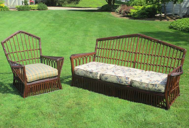 Stick wicker sofa and chair set in dark stained finish with red and black painted trim. High peaked backs and elongated triangular shaped flat arms. Evenly spaced large single reeds overall. Right arm of chair has a woven built-in ash tray holder