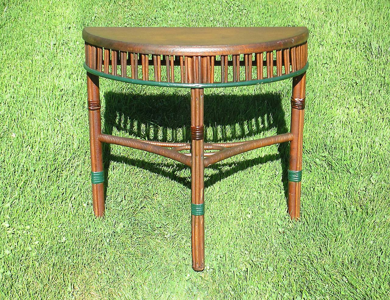 Stick wicker demilune table in natural stained finish with green painted trim. Vertical paired reed apron. Wood top with beveled edge.