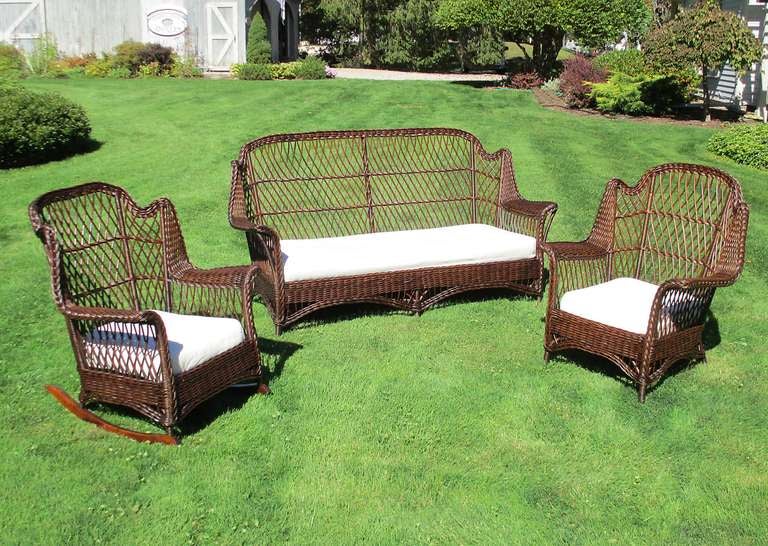 Large scale Bar Harbor style three-piece wicker set in dark natural stained finish. Wing design at shoulders with arched crown backs.  Wide flared arms, cane-wrapped legs.  American Reed & Willow Furniture Co, Wakefield, MA label.  Measurements as