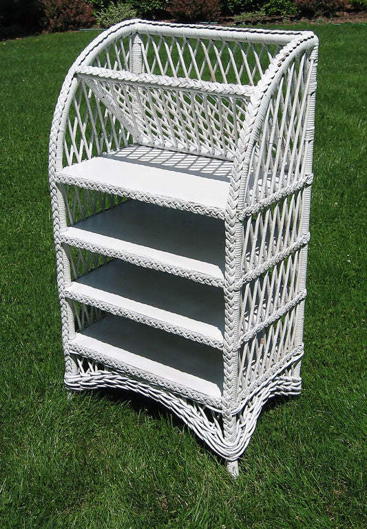 Bar Harbor wicker bookcase in white painted finish. Four solid oak shelves finished with braided border, & angled top shelf  for magazines,etc. Arched sides lead to the high gallery on top. Gently splayed front legs with full skirting. Traditional