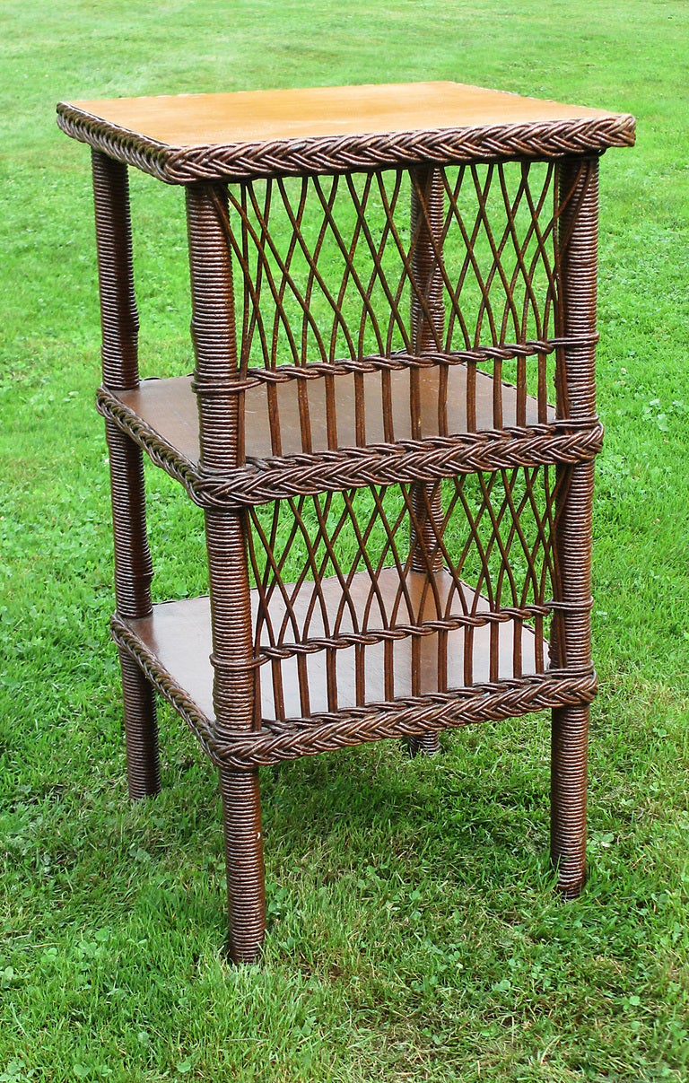 Bar Harbor style square wicker table in natural stained finish. One side with lattice woven panel. A compact table with ample surfaces for storage/display on three wood shelves, each bordered in braiding.