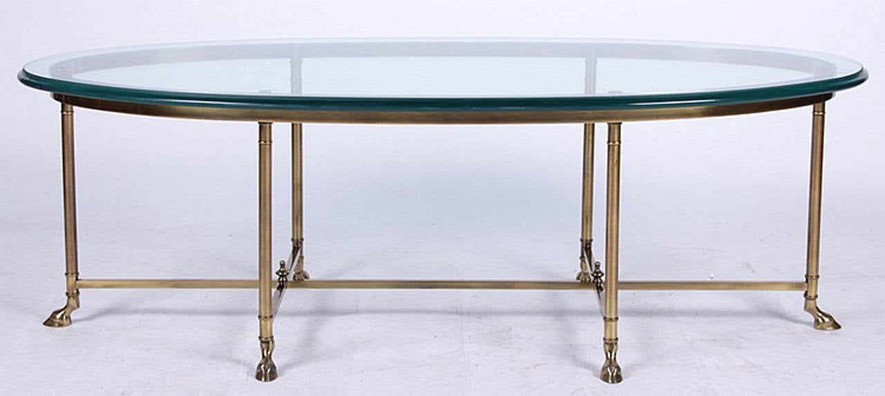 An elegant neoclassical Maison Jansen style brass and glass oval coffee table with six cylindrical legs that end in hoof feet. Glass top has a polished and molded edge.