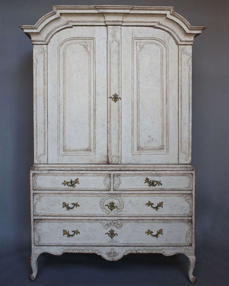 Beautiful rococo cabinet in two parts, Sweden circa 1760. The upper section has three shelves and two drawers behind raised panel doors, all under a bold arched pediment. The lower section has two drawers over two, all with carved detail, and