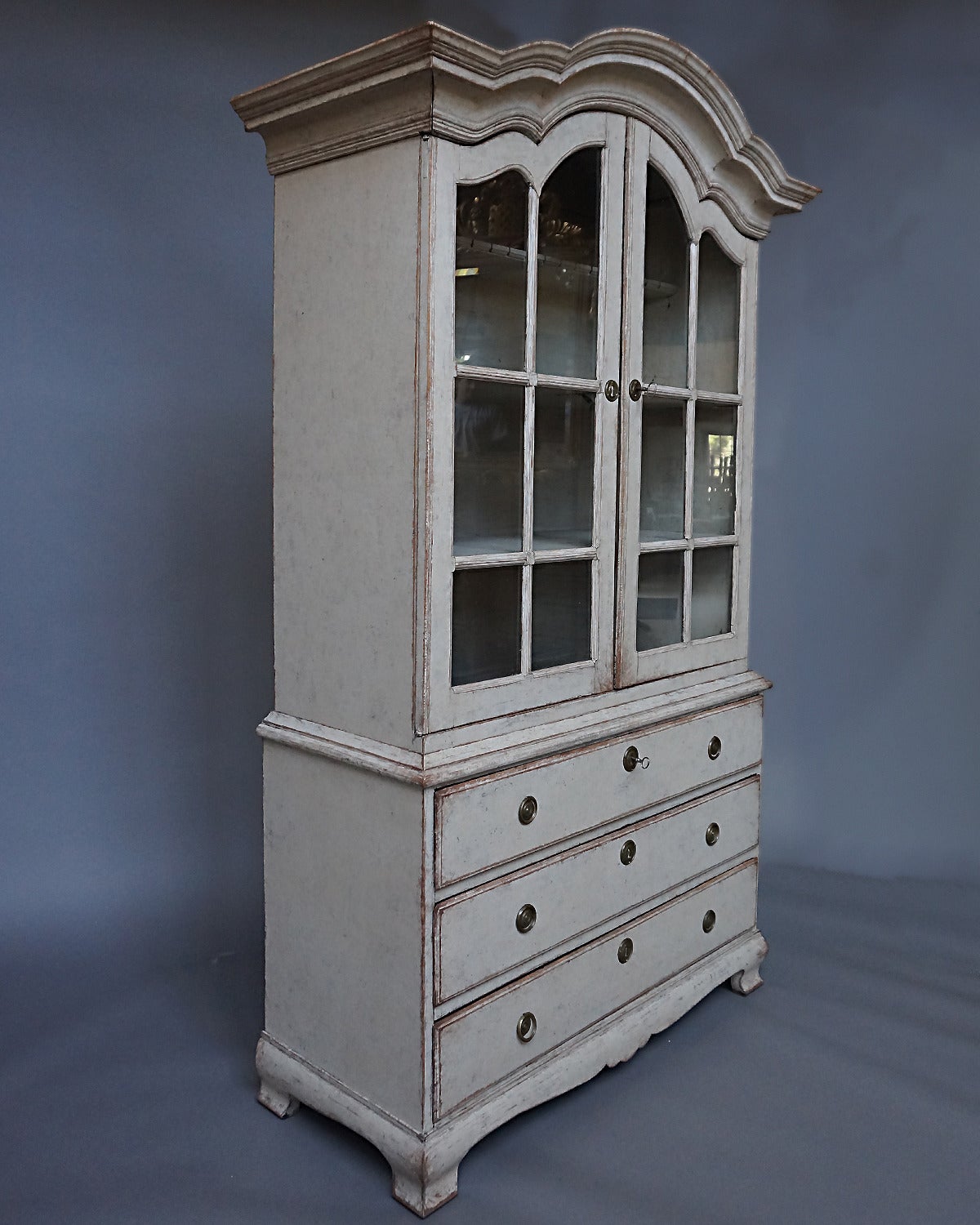 Period Swedish Rococo cabinet, circa 1820, with three shelves behind glazed doors on the upper section and three drawers below. Arched cornice and shaped bracket base, original glass and hardware.