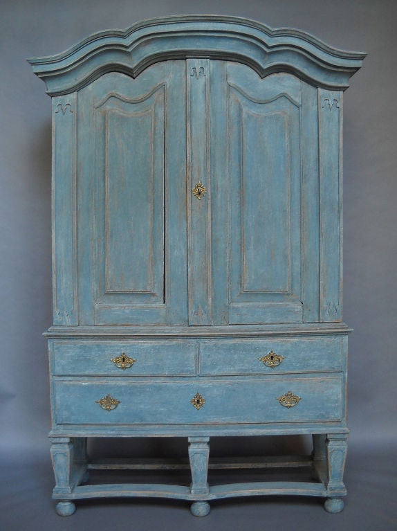 Swedish cabinet in two parts in worn blue paint with original brass fittings. The upper section has two raised panel doors and carved vertical elements under a bonnet cornice. The interior has three shelves and three drawers with brass pulls. The