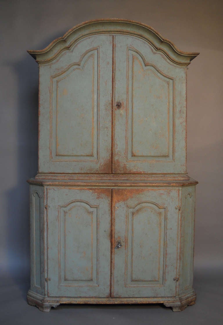 Two part baroque cabinet, Sweden circa 1800, in worn blue paint. The upper section has two raised panel doors under a bold arched cornice and three shelves and four small drawers inside. The lower section has raised panels on the two doors and on
