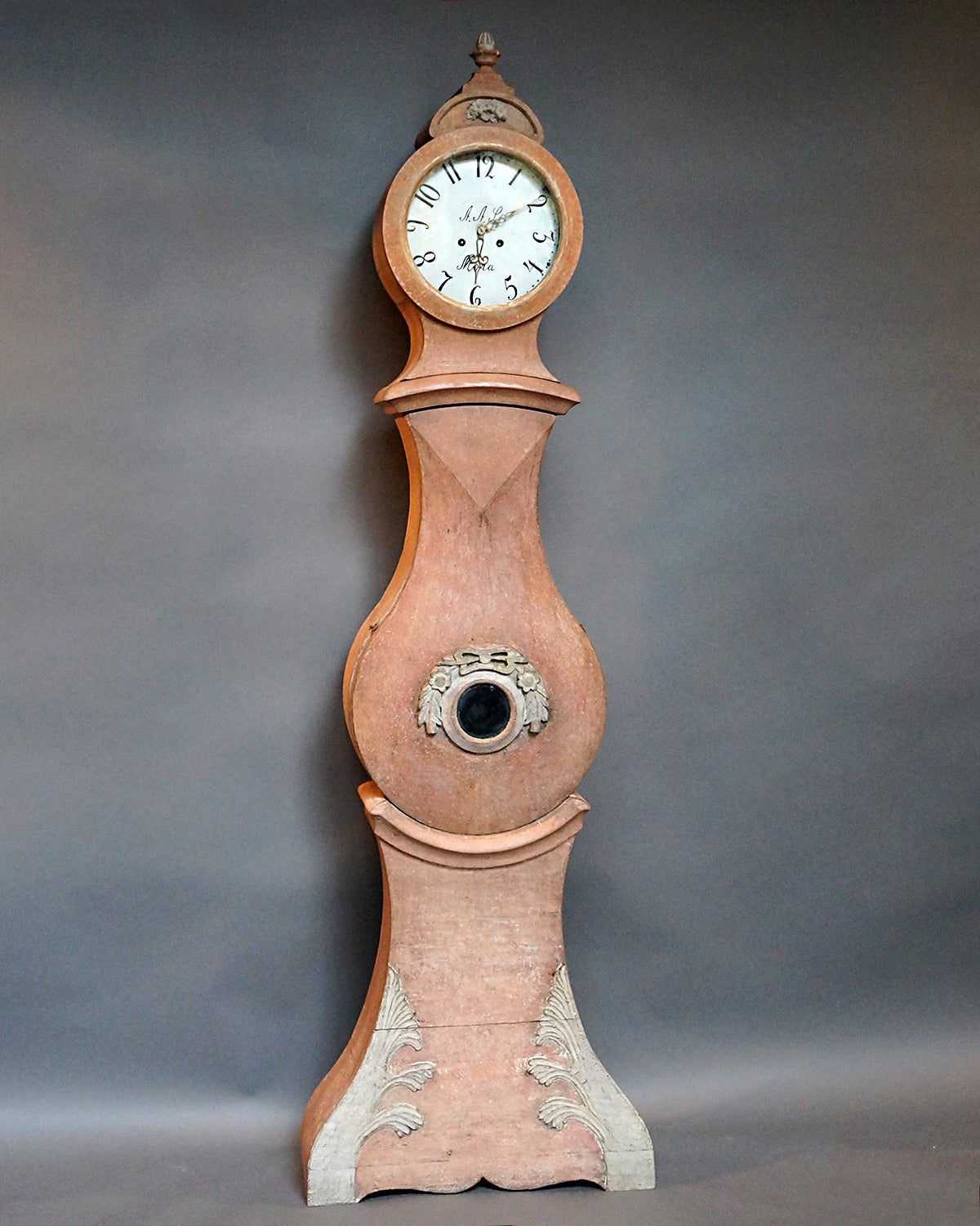 Signed Mora clock, Sweden, circa 1820, in original salmon paint and with its original clock works. The case features an interesting shaped front with applied floral and foliate carving, while the bonnet has a nosegay of flowers and a carved urn with