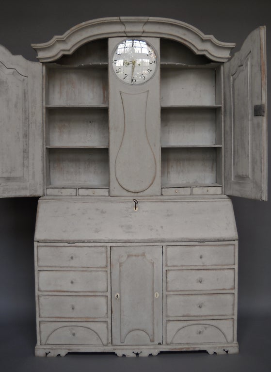 Swedish slant-front secretary, circa 1800, with clock. The upper section has shelves behind raised panel doors on either side of the clock casing and architectural detail on the pediment. The lower section has four banks of three drawers on the