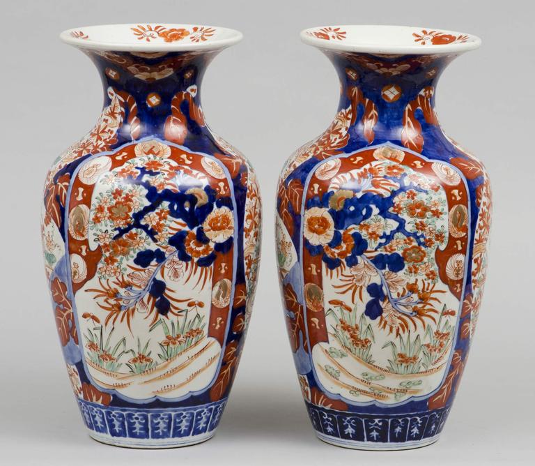 Tall pair of Imari open vases of baluster form with flared necks painted in a palette of iron reds, blue and green with parcel gilding, decorated with opposing  shield-shape panels of blossoms, stems, bold large leaves, berries, all on a dark blue