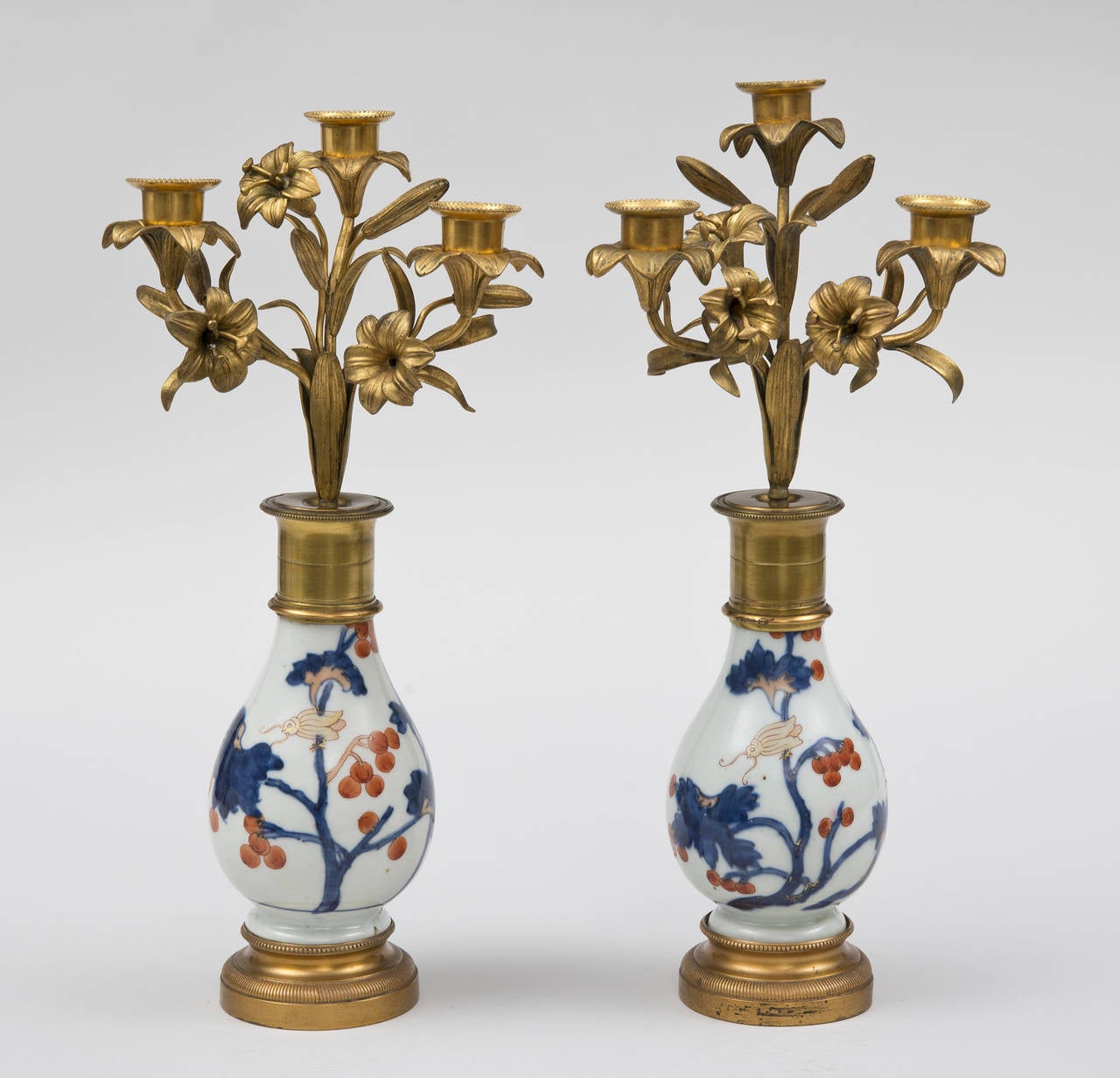 Pair of Louis XVI style gilt metal three-light candelabrum on baluster-shaped “Imari” porcelain stands decorated with blue flowers, red berries and insects; the fruit and foliate stems supporting the lily-form arms. Raised on gilt metal bases.

 