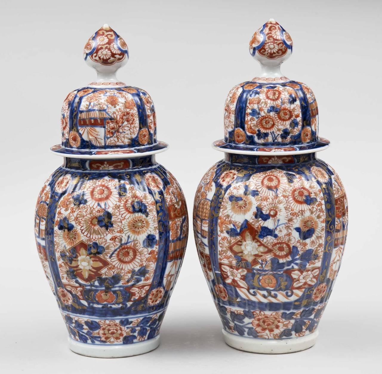 Pair of Imari ribbed vases with covers, the domed and ribbed lid with large finial, four shield-shaped panels on the body decorated with pagodas, trees and flowers in iron red and cobalt blue.

