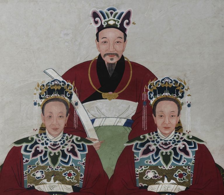 Chinese ancestor portrait depicting three family members, a husband and his two wives.  All wearing red robes with an insignia badge of a white bird and elaborate headdresses. The husband is wearing a gold necklace with a pink jade stone.  Tempera
