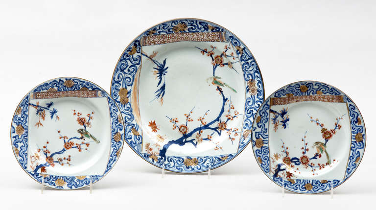 Three Kangxi/Yongzheng plates decorated in underglaze blue, iron red, green black and gold with a border of chrysanthemum blossoms among stylized branches framing a half-open scroll, central scene of a bird perched on a prunus branch opposite a