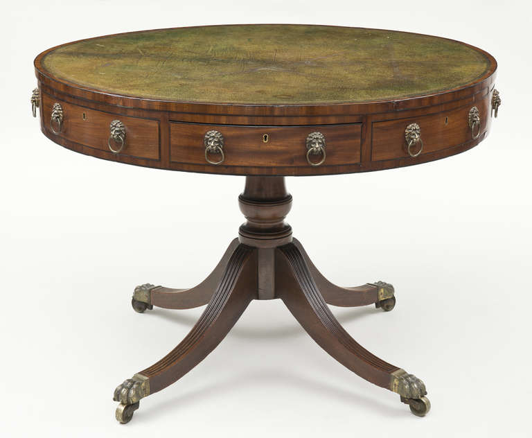 Regency period mahogany revolving drum table with olive green gilt-tooled leather top, the frieze fitted with alternating real and faux drawers, lion head ring pulls, raised on a vase-turned pedestal and four reeded splay legs ending in brass lion