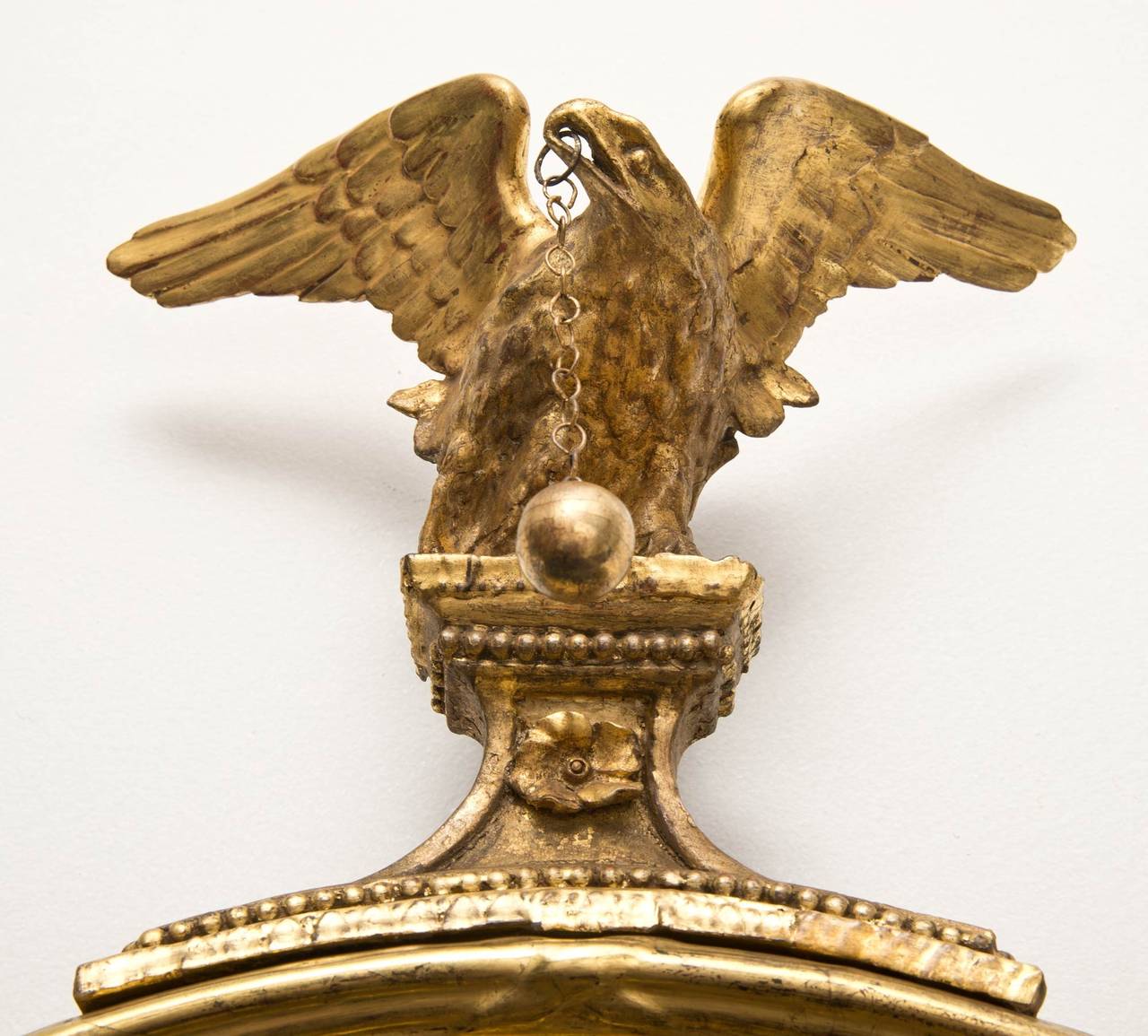 A fine Regency giltwood convex mirror with an ebonized slip, the inner border with tiny beading, the outer border with larger beading, surmounted by an eagle with outspread wings perched on a plinth, a ball and chain hanging from its beak.

Item