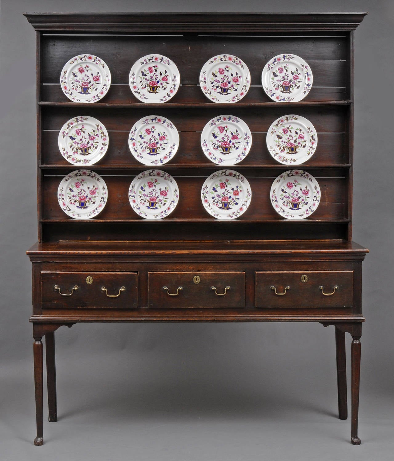 The English oak dresser is in two sections, plate rack and base; base with three drawers, brass swan neck handles, turned delicate front legs ending in pad feet and straight back legs, rack with three shelves and molded cornice. Rack does not have