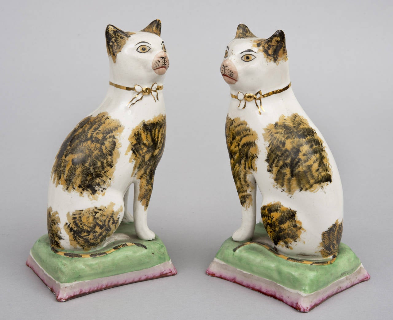 Pair of Staffordshire tabby cats with separate legs, pink nose, gilded ribbon around its neck, black and golden sponge decoration, sitting on a green and pink cushion.

Item #7183